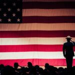 man standing on stage facing an american flag
