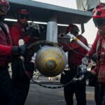 U.S. Navy sailors load a 2,000-pound MK84/BLU-117 bomb onto an F/A-18C Hornet aboard an aircraft carrier.