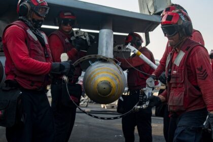 U.S. Navy sailors load a 2,000-pound MK84/BLU-117 bomb onto an F/A-18C Hornet aboard an aircraft carrier.